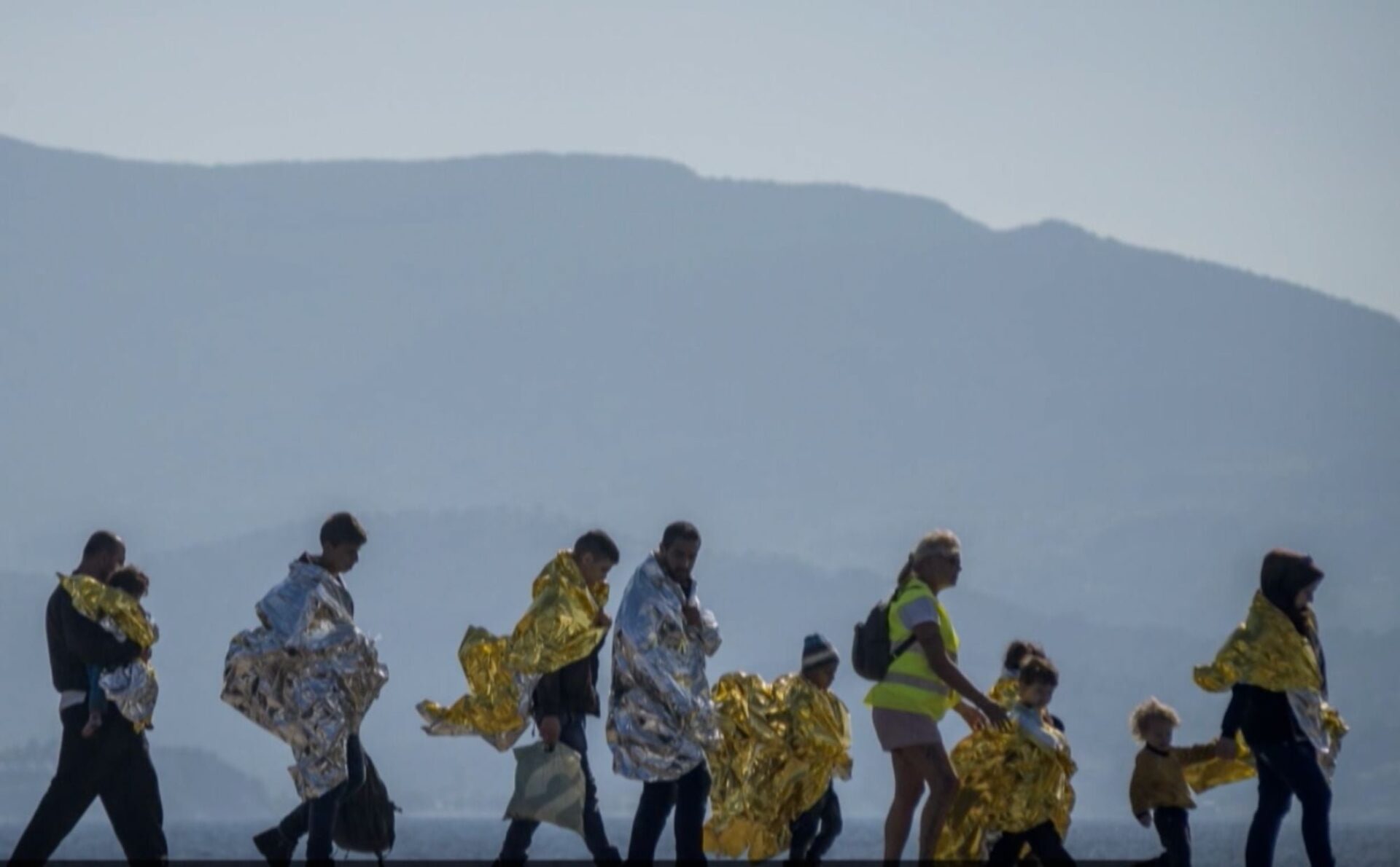 Grupa uchodźców okryta foliami NRC idzie gęsiego u wybrzeży Lampedusy