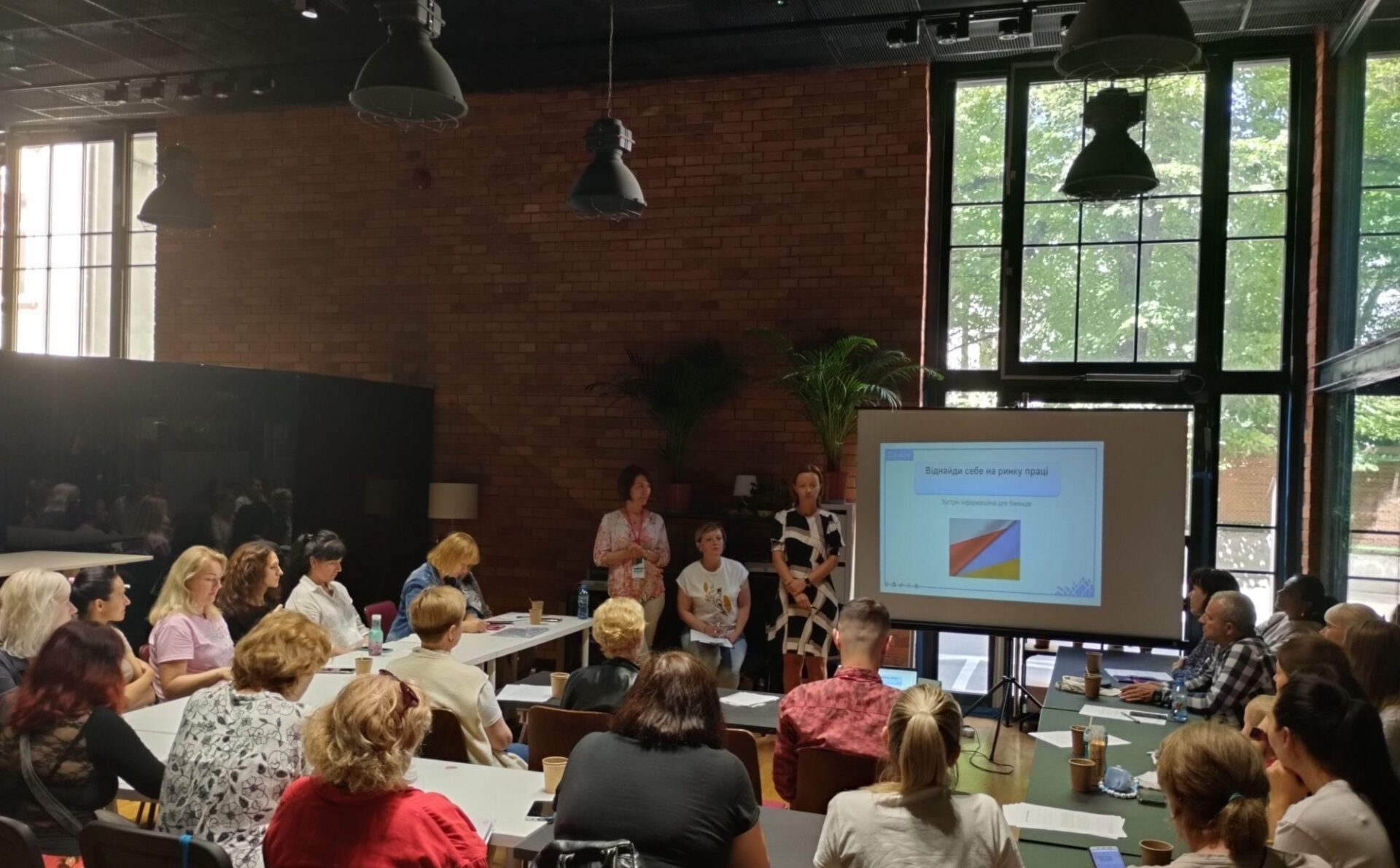 Grupa ludzi w różnym wieku siedzi przy stołach na spotkaniu o projekcie aktywizacji zawodowej. Spotkanie prowadzą trzy kobiety. Grupa zgromadzona jest w sali z dużymi oknami i ścianami z cegły