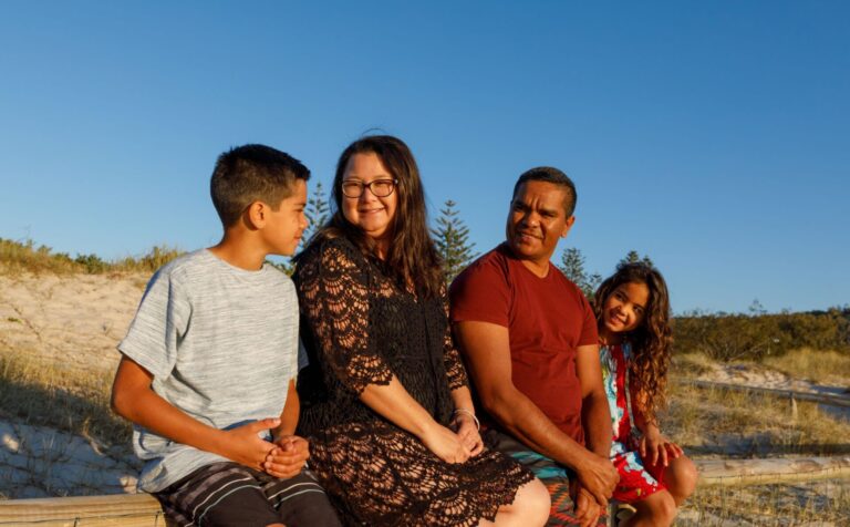 rodzina z Australii: mama, ojciec, syn i córka siedzą gdzieś na zewnątrz. Uśmiechają się. W tle widać niebieskie niebo.