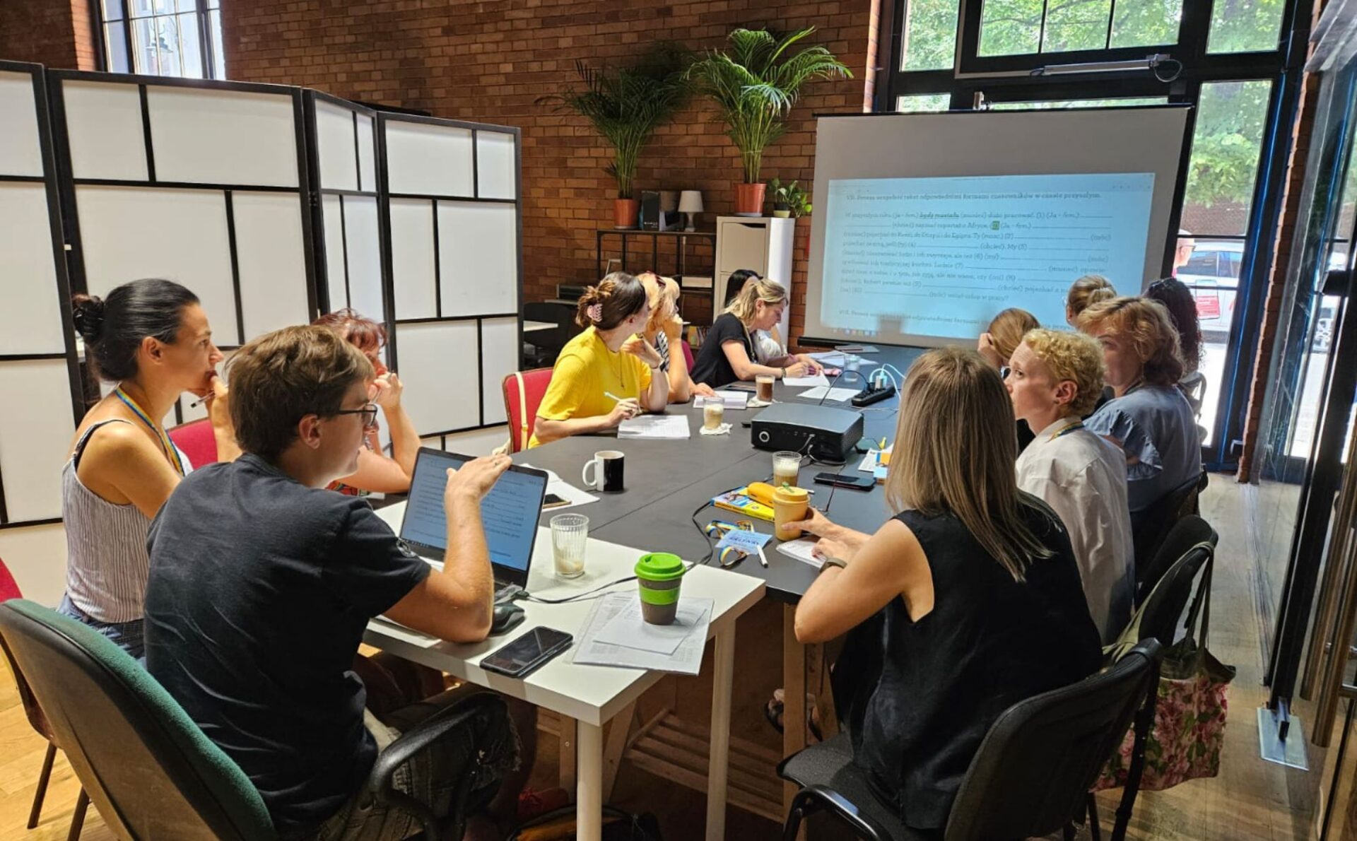 Grupa ludzi w różnym wieku siedzi przy stole i ogląda prezentację