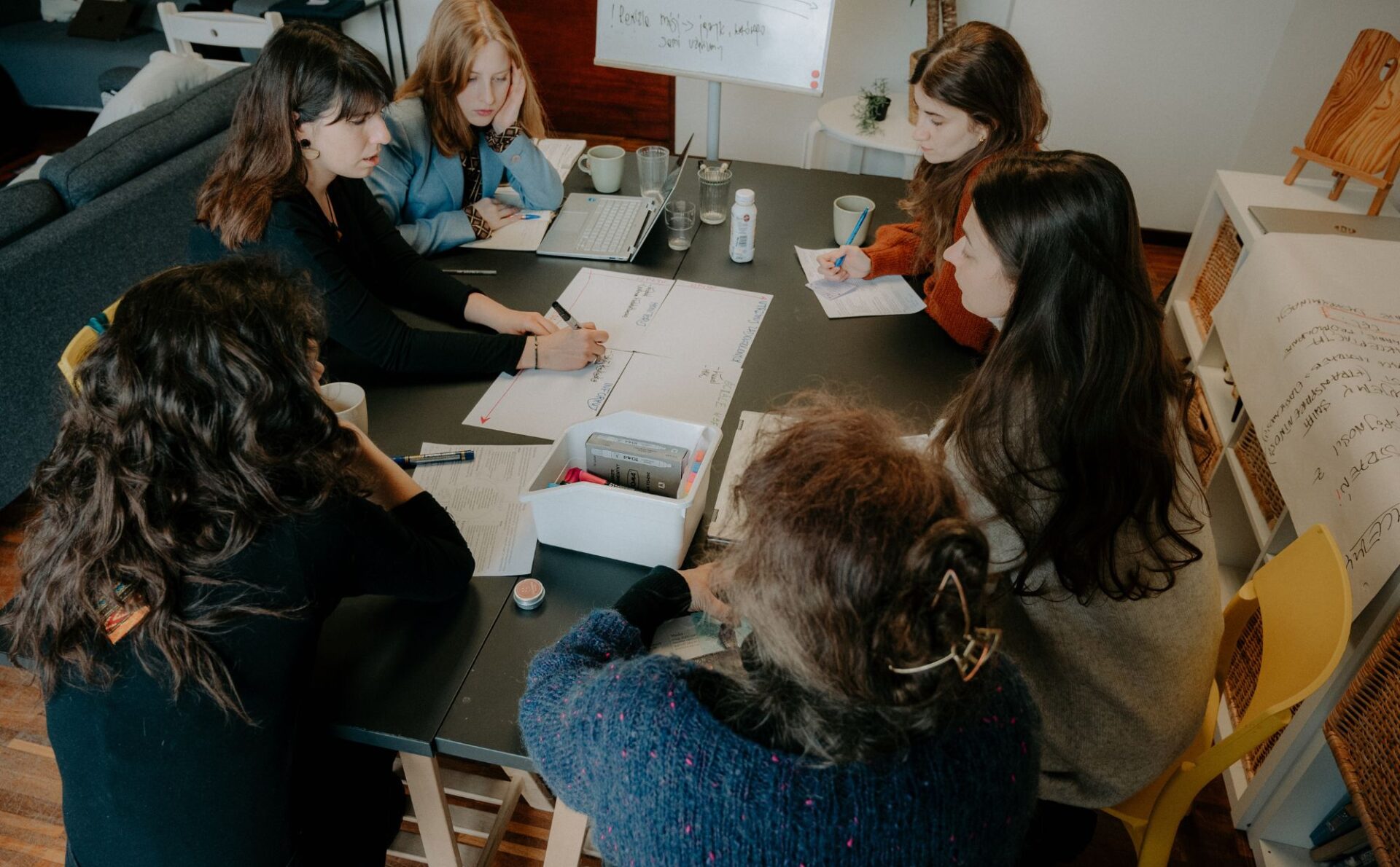 kobiety pracujące nad czymś przy stole, zapisują na kartkach, w tle tablica